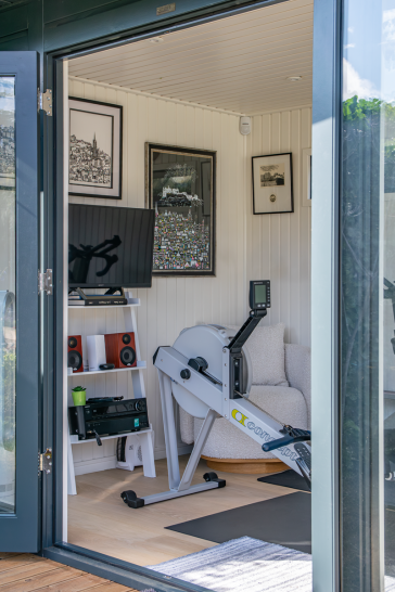 Our Holt Studio as a Home Gym Interior of a compact garden room home gym with a rowing machine, television, speakers and armchair, viewed through open glazed doors.